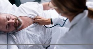 Mann wird im Krankenbett mit Stethoskop abgehört, Credit: Canva