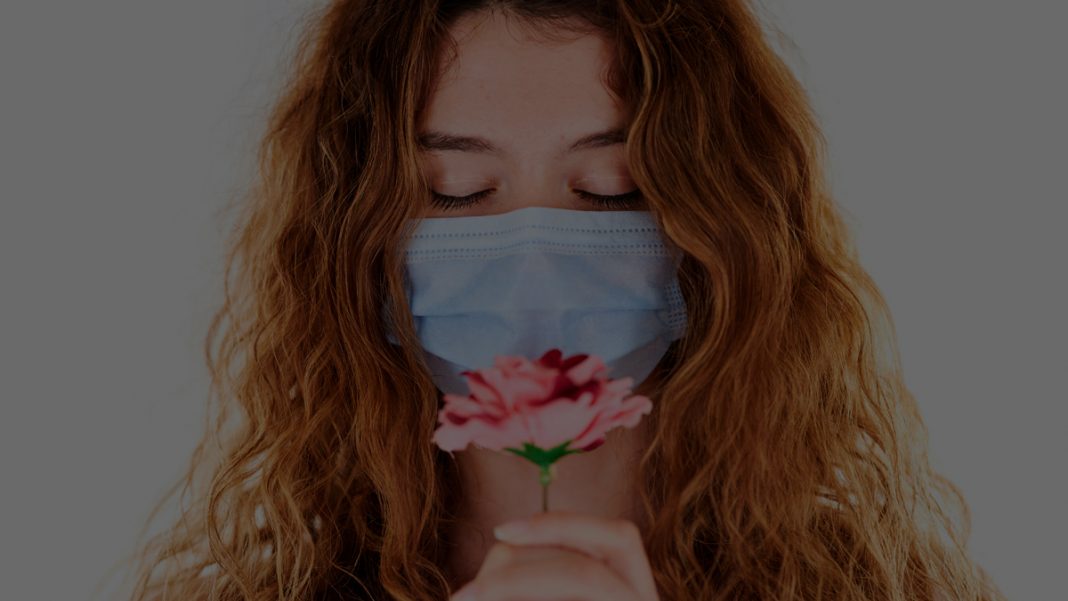 junge Frau mit roten Haaren und Mund-Nasenschutz riecht an Blume, Credit: engin akyurt, Unsplash